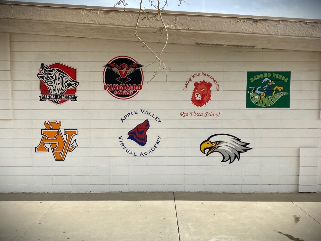 Exterior brick school wall displaying seven different school mascot logos and names as wall graphics.