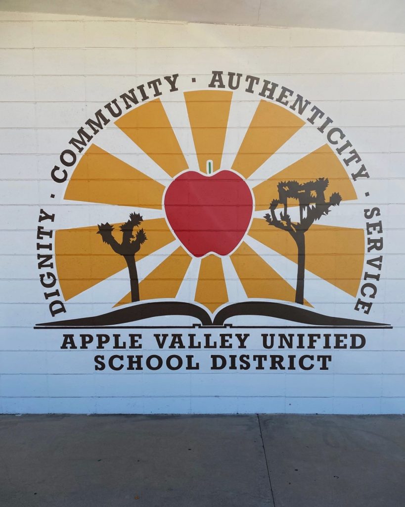 Mural of Apple Valley Unified School District logo with an apple, sun rays, and desert trees on a white brick wall.